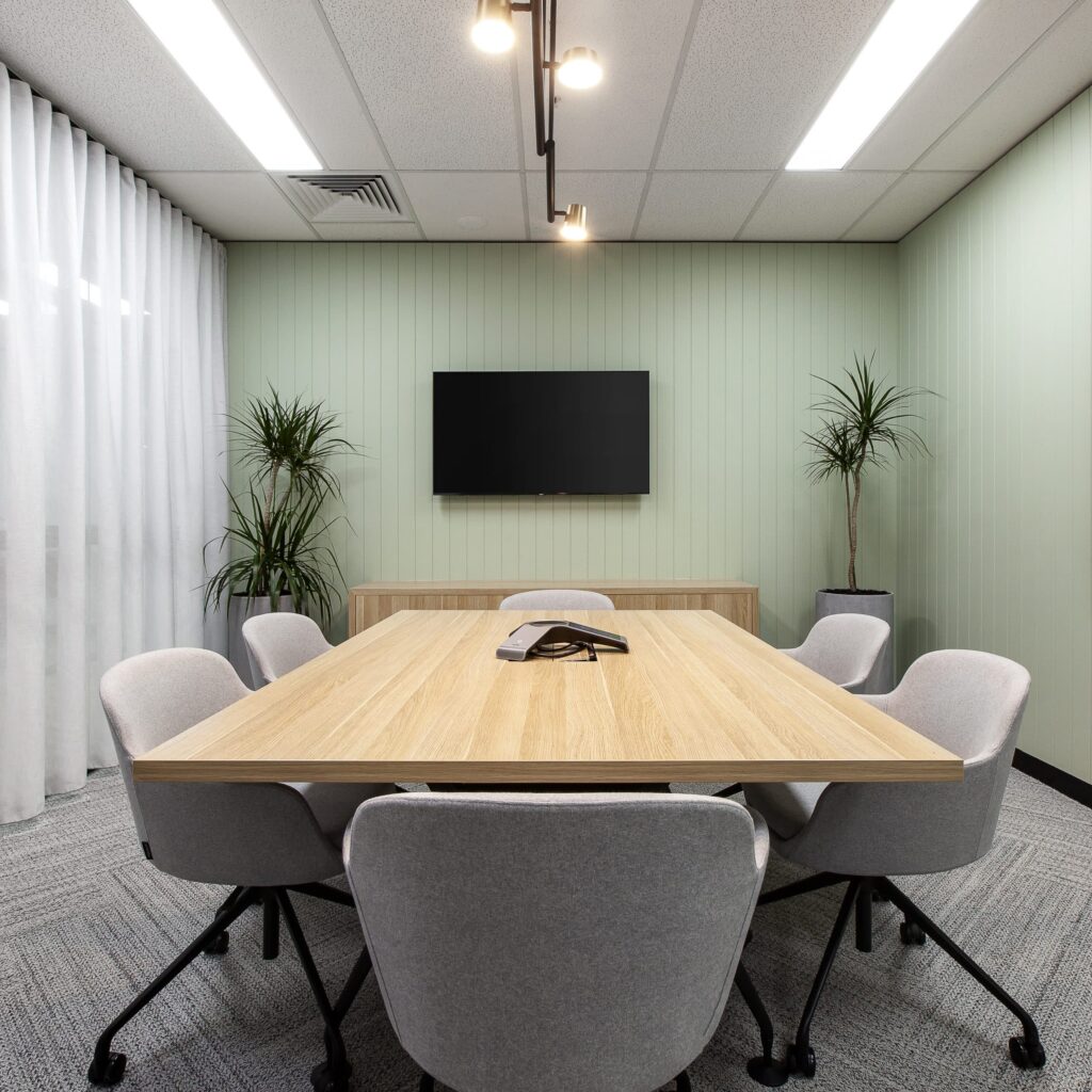 A six seater meeting room with sage green walls and two plants in the corners.