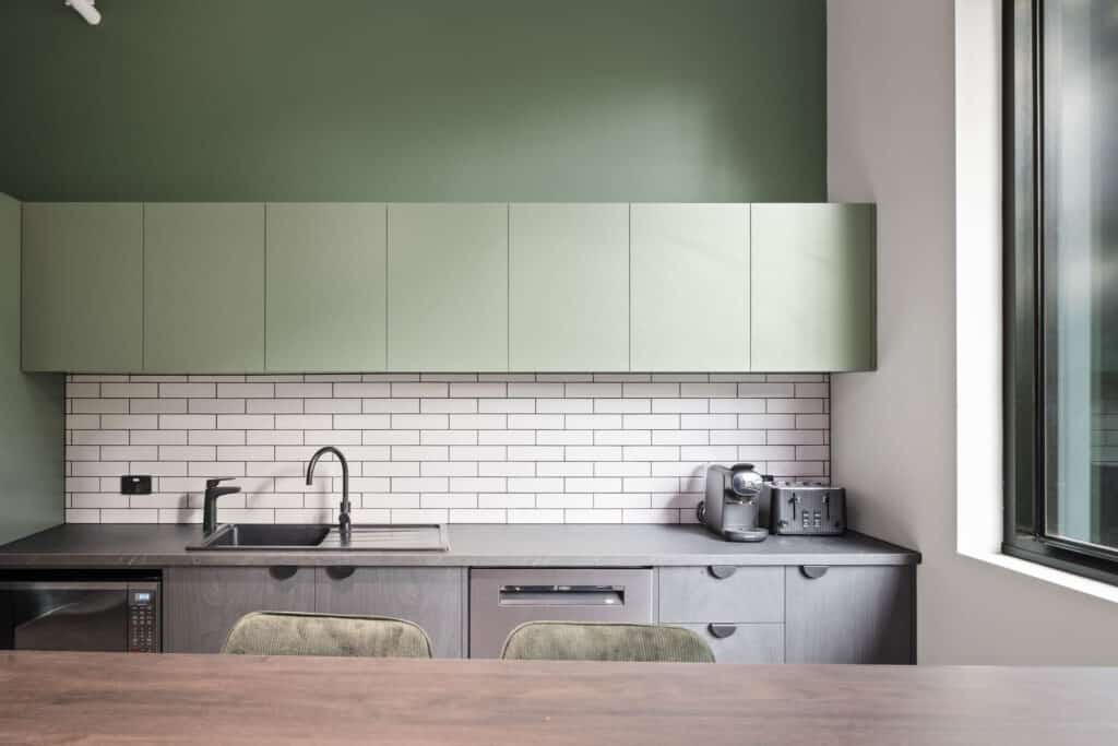 A sage green kitchen space with sink, coffee machine and toaster. A dining table with 2 chairs is in the foreground. 